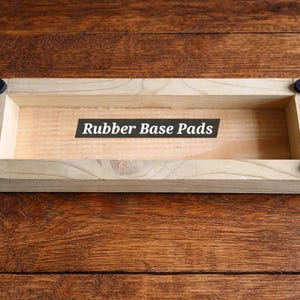 May include: A rectangular wooden tray with rounded corners and black rubber pads. The text "Rubber Base Pads" is printed on a black label inside the tray. The tray is made of light-colored wood.