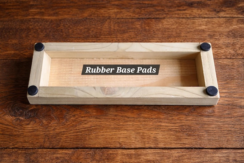 May include: A rectangular wooden tray with rounded corners and black rubber pads. The text "Rubber Base Pads" is printed on a black label inside the tray. The tray is made of light-colored wood.