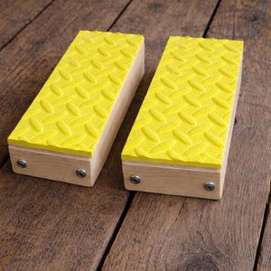 May include: Two wooden blocks with bright yellow, diamond-patterned rubber tops. The blocks are rectangular and have small metal fasteners on the sides. They are set on a rustic, wooden surface.