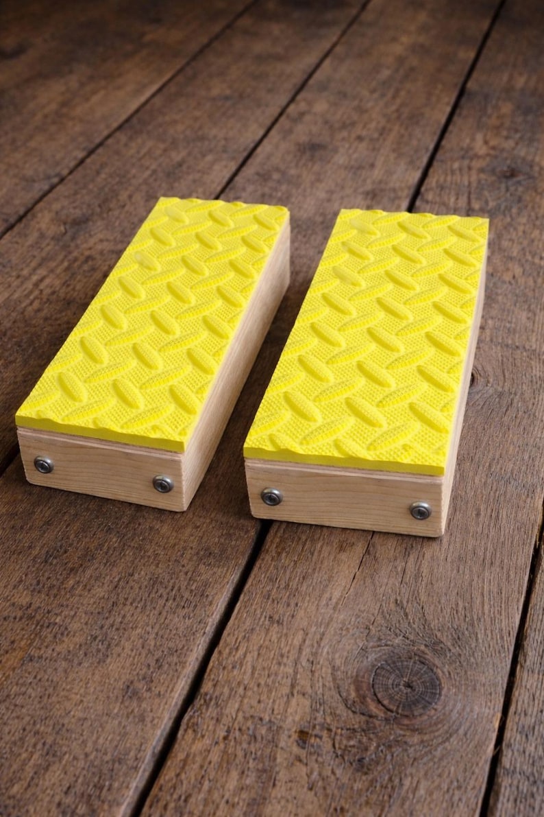 May include: Two wooden blocks with bright yellow, diamond-patterned rubber tops. The blocks are rectangular and have small metal fasteners on the sides. They are set on a rustic, wooden surface.