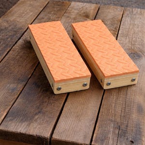 May include: Two rectangular wooden blocks with orange, textured tops. The blocks are made of light-colored wood and are secured with screws. They are resting on a weathered wooden surface.