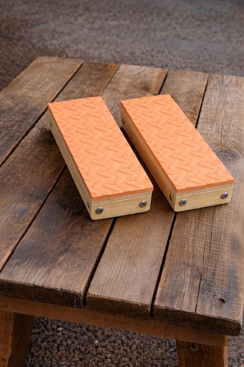 May include: Two rectangular wooden blocks with orange, textured tops. The blocks are made of light-colored wood and are secured with screws. They are resting on a weathered wooden surface.