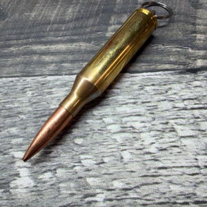 Può includere: Un portachiavi in metallo a forma di proiettile con bossolo in ottone lucido e punta color rame. La parte superiore del bossolo è dotata di un anello metallico argentato per il fissaggio a chiavi o ad altri oggetti di uso personale.