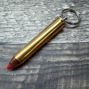 May include: A metallic bullet-shaped keyring featuring a polished brass casing, a copper-coloured projectile tip, and a bright red polymer insert at the very point. A silver-toned metal split ring is attached to the base of the brass casing.