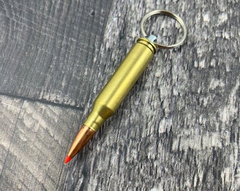 Könnte beinhalten: Ein goldfarbener Schlüsselanhänger in Form einer Patronenhülse mit einem silbernen Schlüsselring. Die Patronenhülse hat eine rote Spitze.