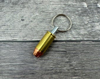 Könnte beinhalten: Ein goldfarbener Patronenhülsen-Schlüsselanhänger mit einem silbernen Ring. Die Patronenhülse hat eine kupferne Spitze.