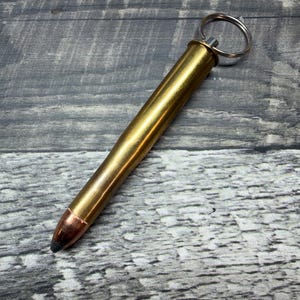 Op de afbeelding: Een messingkleurige metalen pen in de vorm van een kogelpatroon van groot kaliber. Het schrijfinstrument heeft een koperkleurige punt en een zilverkleurige metalen sleutelring aan het platte uiteinde, waardoor het als sleutelhanger kan dienen.