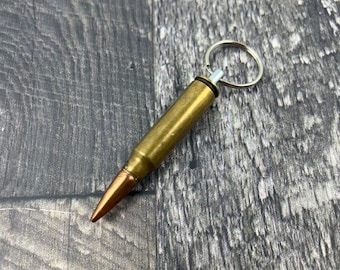 May include: A gold-colored bullet casing keychain with a silver key ring. The keychain is on a dark wood surface.