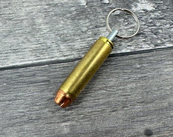 Könnte beinhalten: Ein goldfarbener Schlüsselanhänger in Form einer Patronenhülse mit einem silbernen Schlüsselring. Die Patronenhülse hat eine kupferfarbene Spitze.