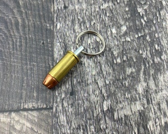 Könnte beinhalten: Ein goldfarbener Schlüsselanhänger in Form einer Patronenhülse mit einem silbernen Ring. Der Schlüsselanhänger liegt auf einer Holzoberfläche.