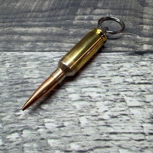 Puede incluir: Un llavero que presenta un casquillo de bala metálico con un cuerpo cilíndrico de color latón y una punta de color cobre. Un anillo circular de metal plateado está unido a la base del casquillo, lo que permite sujetar las llaves de forma segura.