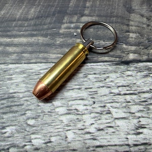 May include: A keychain featuring a brass-colored metal bullet casing with a copper-toned hollow point tip. A circular silver-toned metal split ring is attached to the base of the casing, allowing for the attachment of keys or other small items.