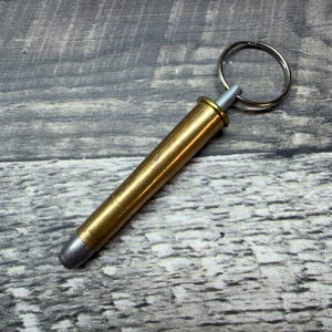 May include: A brass-colored keychain accessory with a silver tip and a silver keyring. The cylindrical body has a gold finish. The item is lying on a weathered, gray wooden surface.