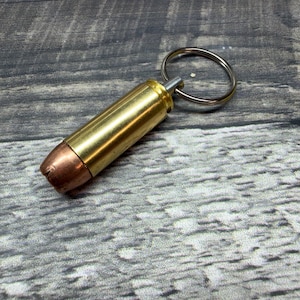Op de afbeelding: Een messing kogel sleutelhanger met een koperkleurige punt en een zilveren sleutelring. De kogel is cilindrisch en heeft een gepolijste afwerking. De sleutelhanger is ontworpen als een uniek accessoire.
