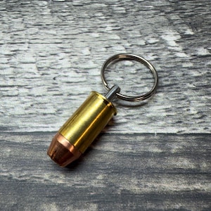 May include: A keychain featuring a metallic bullet-shaped charm with a polished brass casing and a copper-colored projectile tip. The charm is attached to a circular silver-toned metal split ring, providing a functional way to organize keys.
