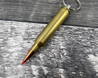 Könnte beinhalten: Ein goldfarbener Patronenhülsen-Schlüsselanhänger mit einem silbernen Ring. Die Patronenhülse hat eine rote Spitze.