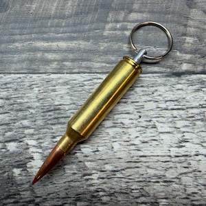 May include: A brass bullet keychain with a silver key ring. The bullet has a gold casing and a copper-colored tip. The keychain is set against a gray wooden background.