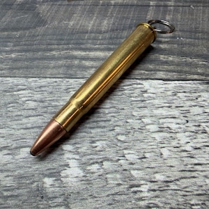 Può includere: Un portachiavi metallico a forma di proiettile con un bossolo in ottone lucido e una punta del proiettile color rame. L'oggetto include un piccolo anello in metallo color argento fissato alla base, progettato per agganciare chiavi o piccoli oggetti.