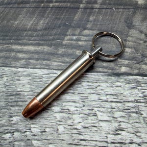 Puede incluir: Un llavero plateado con forma de bala y punta cobriza. El llavero tiene un anillo plateado para sujetar llaves. El artículo está sobre un fondo de madera gris.