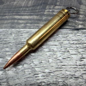 Può includere: Un portachiavi in metallo color ottone a forma di proiettile, dotato di una punta color rame e di un anello metallico argentato fissato all'estremità piatta. L'oggetto presenta una finitura liscia e lucida e un corpo cilindrico che si restringe.
