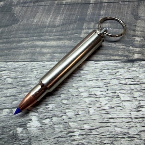 May include: A silver bullet-shaped keyring with a copper tip and a blue point. The keyring has a silver ring attached to the top. The background is a grey wooden surface.