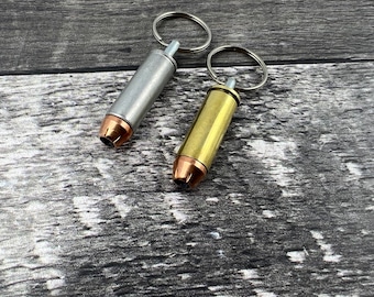 Könnte beinhalten: Zwei Schlüsselanhänger in Form von Patronenhülsen, einer silberfarben und einer golden, mit Schlüsselringen. Die Schlüsselanhänger liegen auf einer grauen Holzoberfläche.