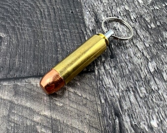 Könnte beinhalten: Ein goldfarbener Schlüsselanhänger in Form einer Patronenhülse mit einem silbernen Ring. Die Hülse hat eine kupferfarbene Spitze.