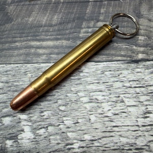 Könnte beinhalten: Ein Schlüsselanhänger aus einer Messing-Patronenhülse mit einer kupferfarbenen Geschossspitze. Der zylindrische Messingkörper hat eine metallische Oberfläche und ist oben mit einem silberfarbenen Metallring zur Befestigung von Schlüsseln versehen.