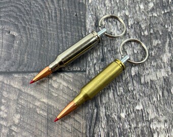 May include: Two bullet casing keychains, one silver and one gold, with red tips and silver key rings. The keychains are on a wooden surface.