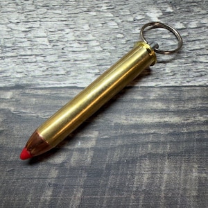 May include: A brass-coloured metal keyring shaped like a rifle cartridge. The object features a polished cylindrical body, a copper-toned bullet tip with a small red plastic insert at the very point, and a circular silver-coloured metal split ring attached.