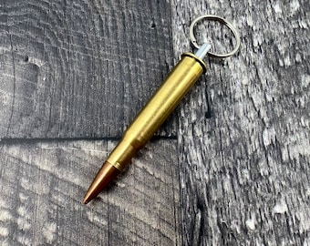May include: A brass bullet-shaped keychain with a silver key ring. The bullet has a copper-colored tip and a gold-colored casing. The keychain is shown against a dark gray wood grain background.