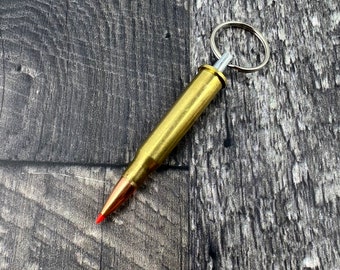 Könnte beinhalten: Ein goldfarbener Schlüsselanhänger in Form einer Gewehrpatrone mit einem silbernen Ring. Die Gewehrpatrone hat eine rote Spitze.