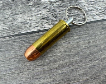 Könnte beinhalten: Ein Schlüsselanhänger in Form einer goldfarbenen Patronenhülse mit einem silbernen Schlüsselring. Die Patronenhülse hat eine kupferfarbene Spitze.
