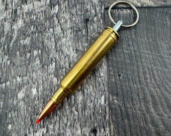 Könnte beinhalten: Ein goldfarbener Schlüsselanhänger in Form einer Patronenhülse mit einem silbernen Schlüsselring. Die Hülse hat eine rote Spitze und liegt auf einem dunklen Holzgrund.