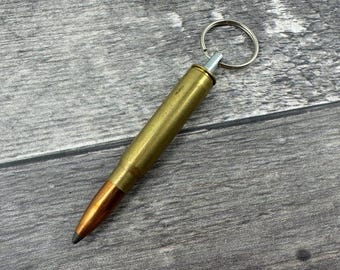 Könnte beinhalten: Ein goldfarbener Schlüsselanhänger in Form einer Gewehrpatrone mit einem silbernen Ring. Der Schlüsselanhänger liegt auf einer grauen Holzoberfläche.