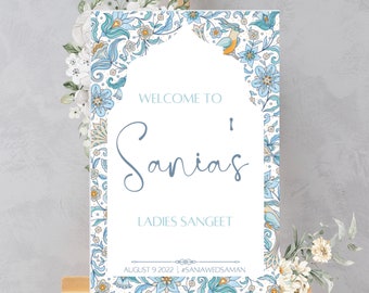 May include: A welcome sign with a blue and white floral design. The sign says "Welcome to Sania's Ladies Sangeet August 9, 2022 #SaniaWedsAman".