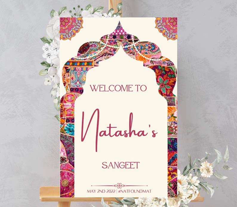 May include: Welcome to Natasha's Sangeet sign with a colorful patterned archway design. The sign is on a wooden easel and has the date May 2nd 2022 and the hashtag #NATIFOUNDMAT.