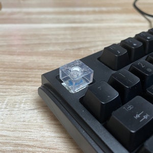 Könnte beinhalten: Eine transparente, aus Kunststoff gefertigte Tastaturkappe mit einem runden, weißen Knopf in der Mitte. Die Kappe liegt auf einer schwarzen Tastatur.
