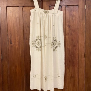 May include: A white linen dress with green embroidered geometric patterns. The dress has a square neckline and thin straps.