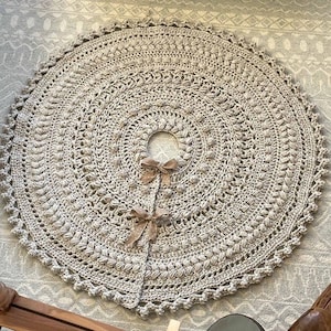 May include: A crocheted beige tree skirt with a scalloped edge and a center hole for a tree trunk. The skirt is decorated with a repeating pattern of small, raised stitches.