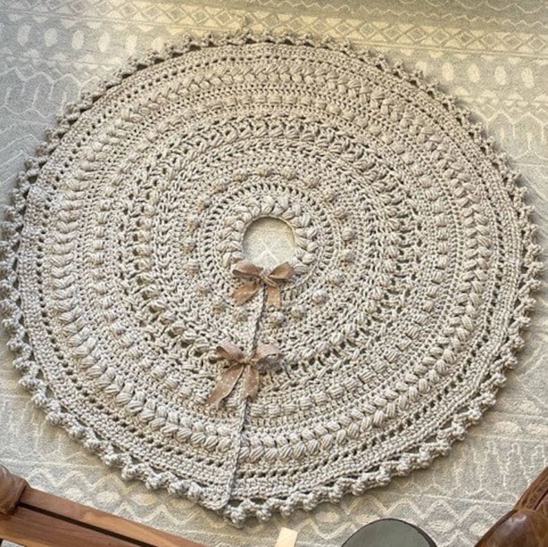 May include: A crocheted beige tree skirt with a scalloped edge and a center hole for a tree trunk. The skirt is decorated with a repeating pattern of small, raised stitches.
