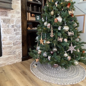 May include: A gray crocheted tree skirt with a white crocheted border surrounds a decorated Christmas tree with ornaments and a variety of decorations. The tree is in front of a fireplace and a bookshelf.