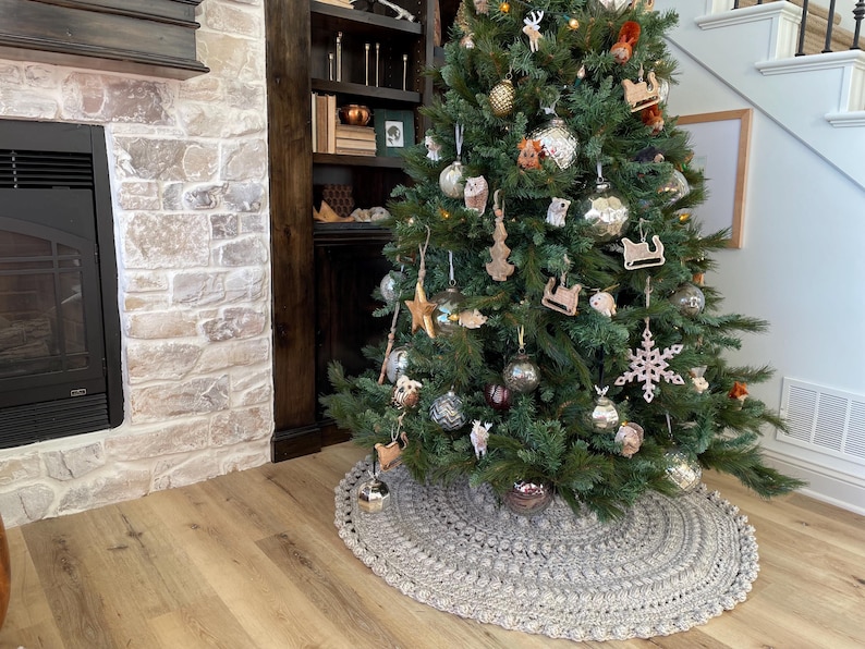 May include: A gray crocheted tree skirt with a white crocheted border surrounds a decorated Christmas tree with ornaments and a variety of decorations. The tree is in front of a fireplace and a bookshelf.