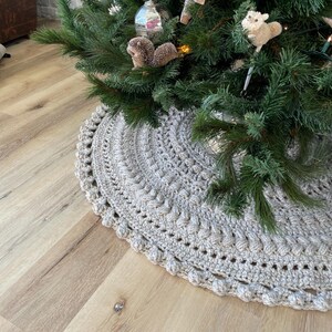 May include: A crocheted beige tree skirt with a textured design and a beaded edge. The skirt is placed on a light wood floor.