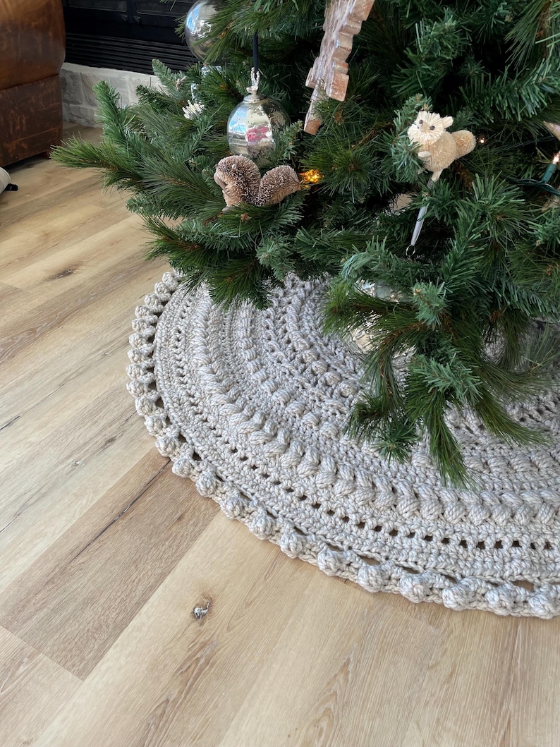 May include: A crocheted beige tree skirt with a textured design and a beaded edge. The skirt is placed on a light wood floor.
