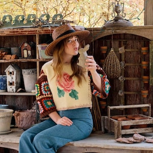 May include: A woman wearing a crocheted sweater with a floral design, a wide-brimmed hat, and blue jeans. She is holding a small ceramic rabbit figurine.