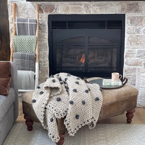May include: A gray and white crocheted throw blanket with black polka dots is draped over a brown leather ottoman with wooden legs. The ottoman is in front of a fireplace with a black metal surround and a roaring fire.
