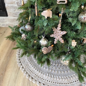 May include: A beige crocheted Christmas tree skirt with a scalloped edge. The skirt is placed on a wood floor and a decorated Christmas tree is in the background.