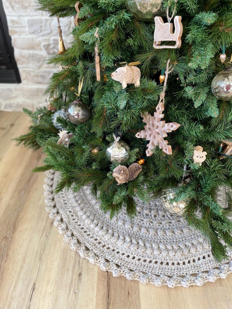 May include: A beige crocheted Christmas tree skirt with a scalloped edge. The skirt is placed on a wood floor and a decorated Christmas tree is in the background.