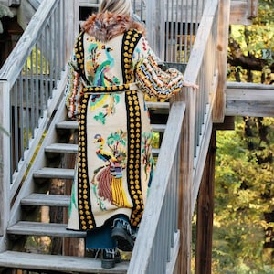 May include: A woman wearing a long, white cardigan with a colorful embroidered design of peacocks and flowers. The cardigan has a black and yellow trim and a fur collar. She is walking down a wooden staircase.